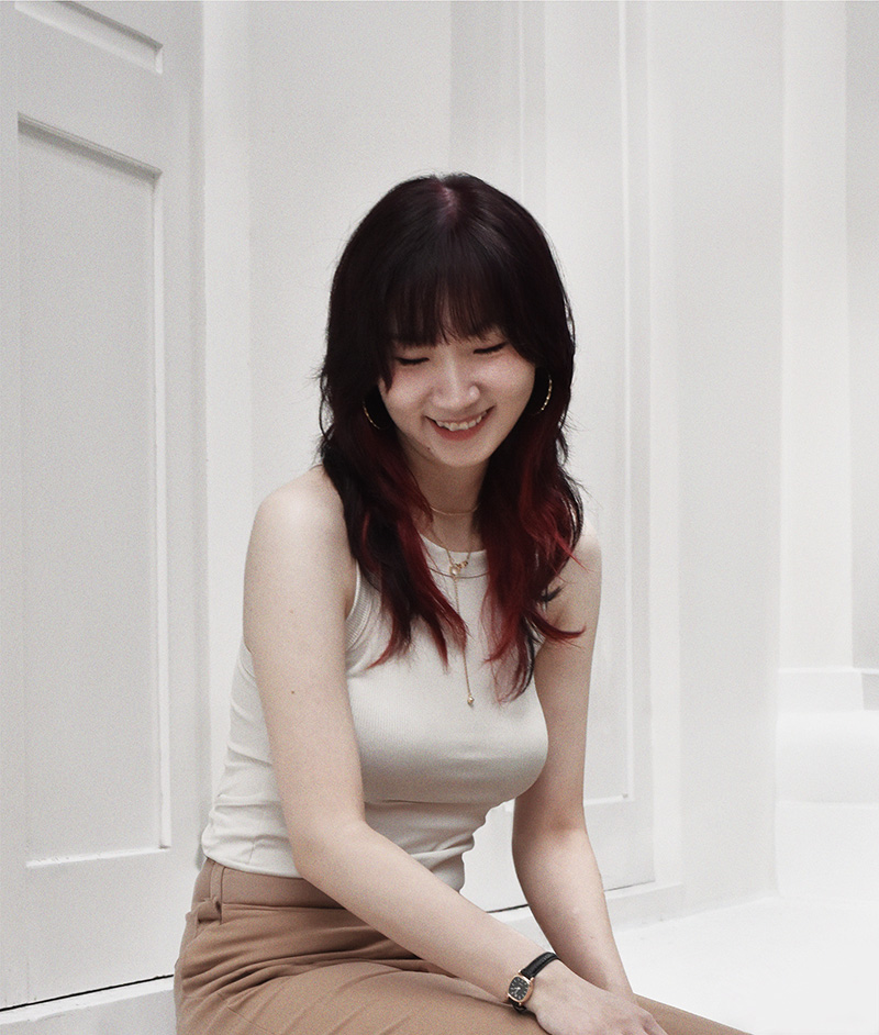 Candid photo of a young Chinese woman with black hair and red streaks, smiling and looking down, wearing a beige tank top against a white panel background during a casual photoshoot.
