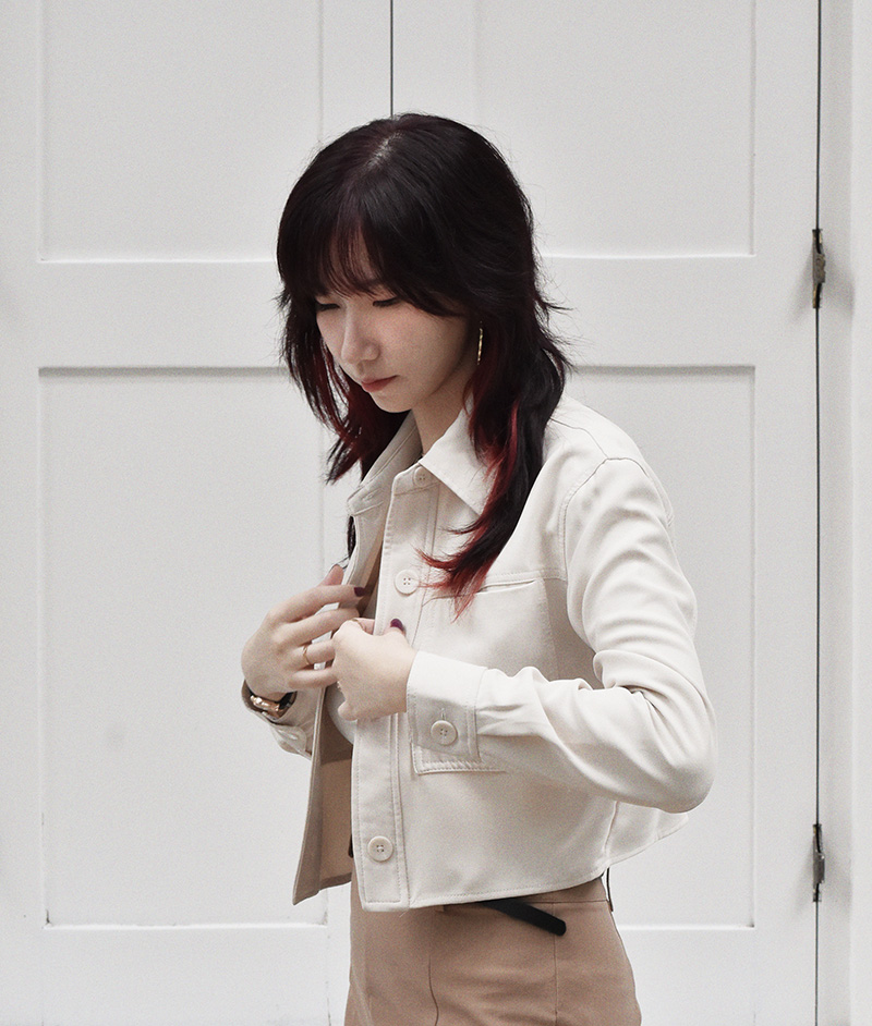 Candid photo of a young Chinese woman with black hair and red streaks, adjusting her beige cropped jacket against a white panel background during a casual photoshoot.
